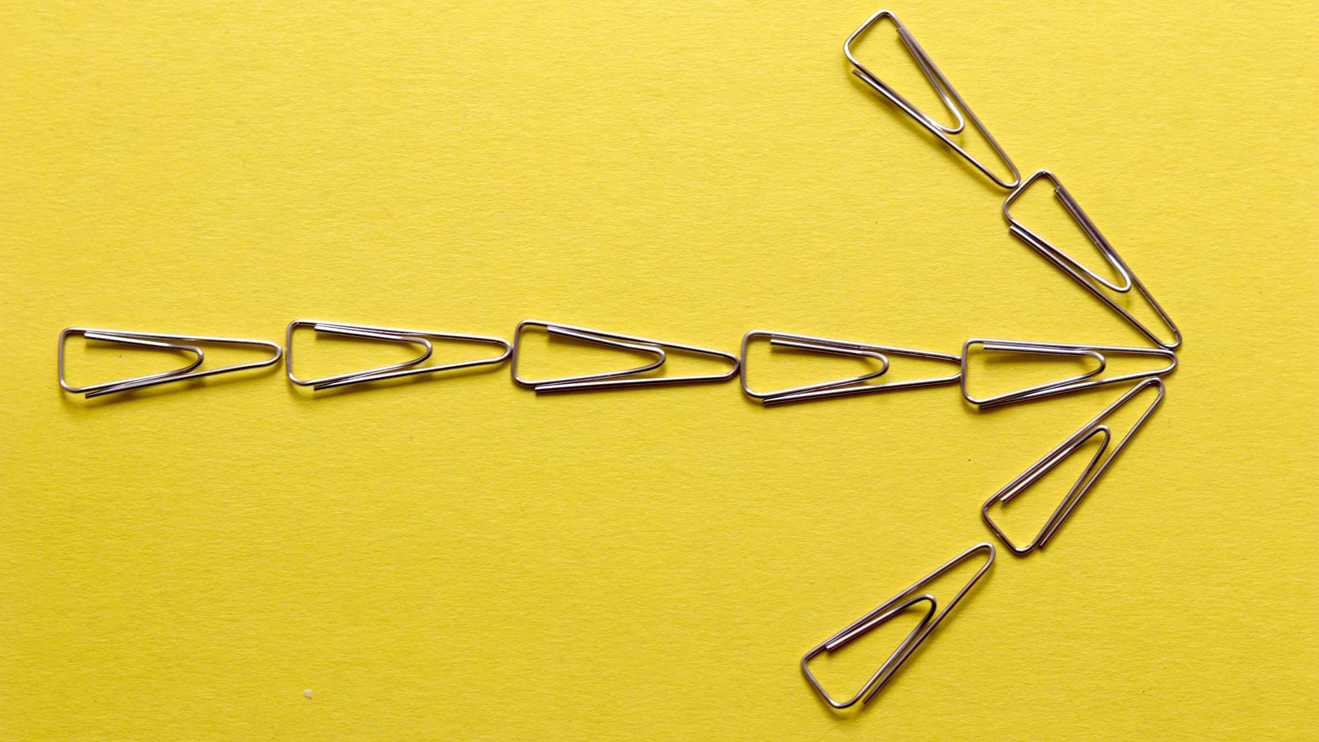 Arrow pointing to the right against a yellow background. The arrow is made of paper clips.