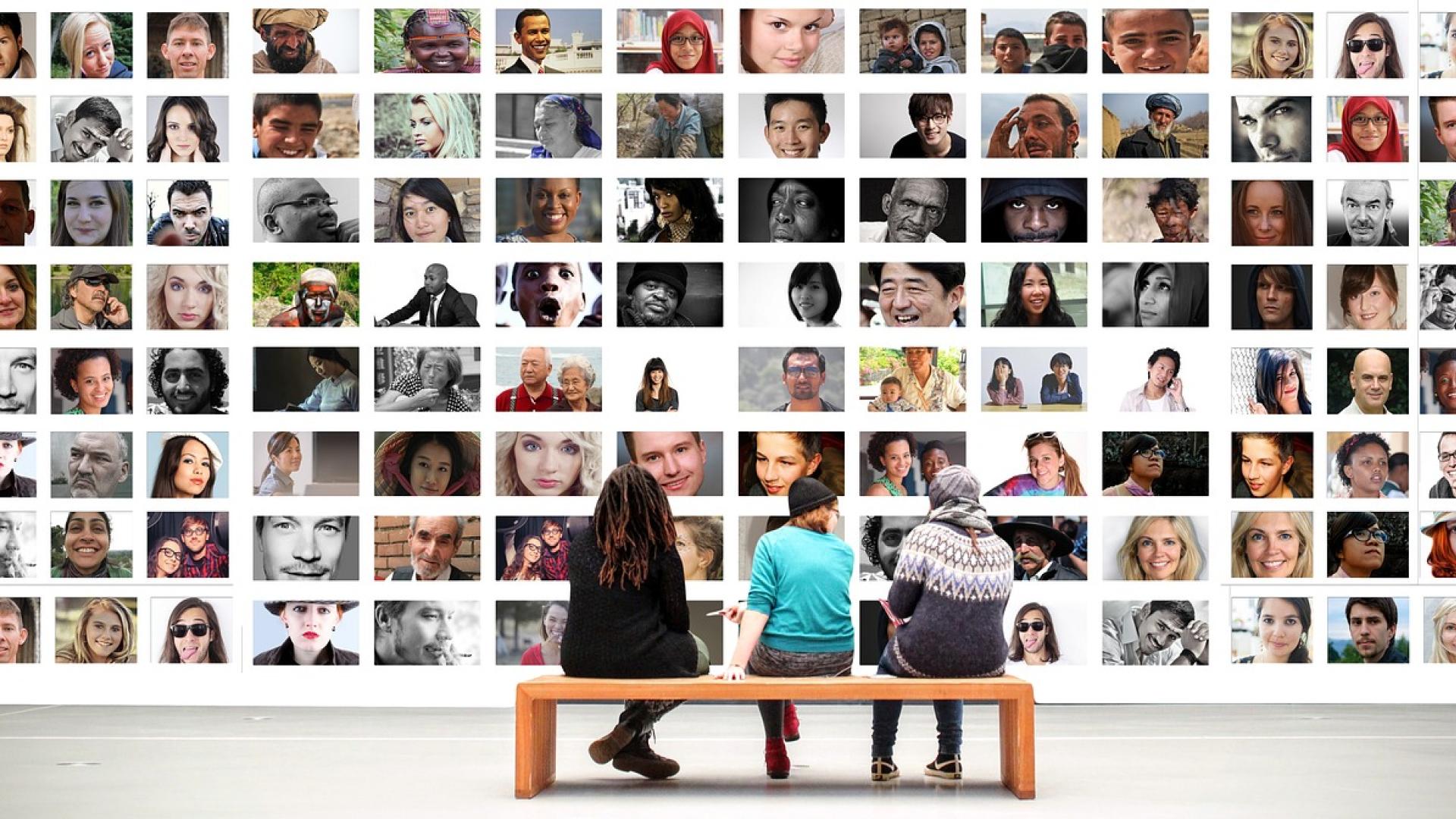 Photo from behind of three people sitting on an art gallery bench viewing a large wall with numerous photos of people