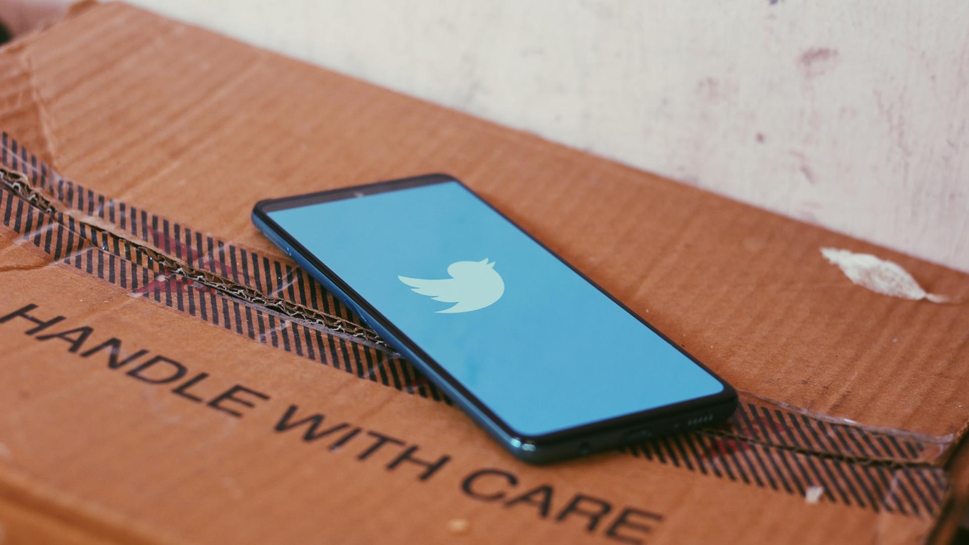 The twitter logo on a phone, on top of a cardboard box saying handle with care